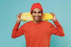 Smiling teen with red hat and sweater carrying a skateboard. 