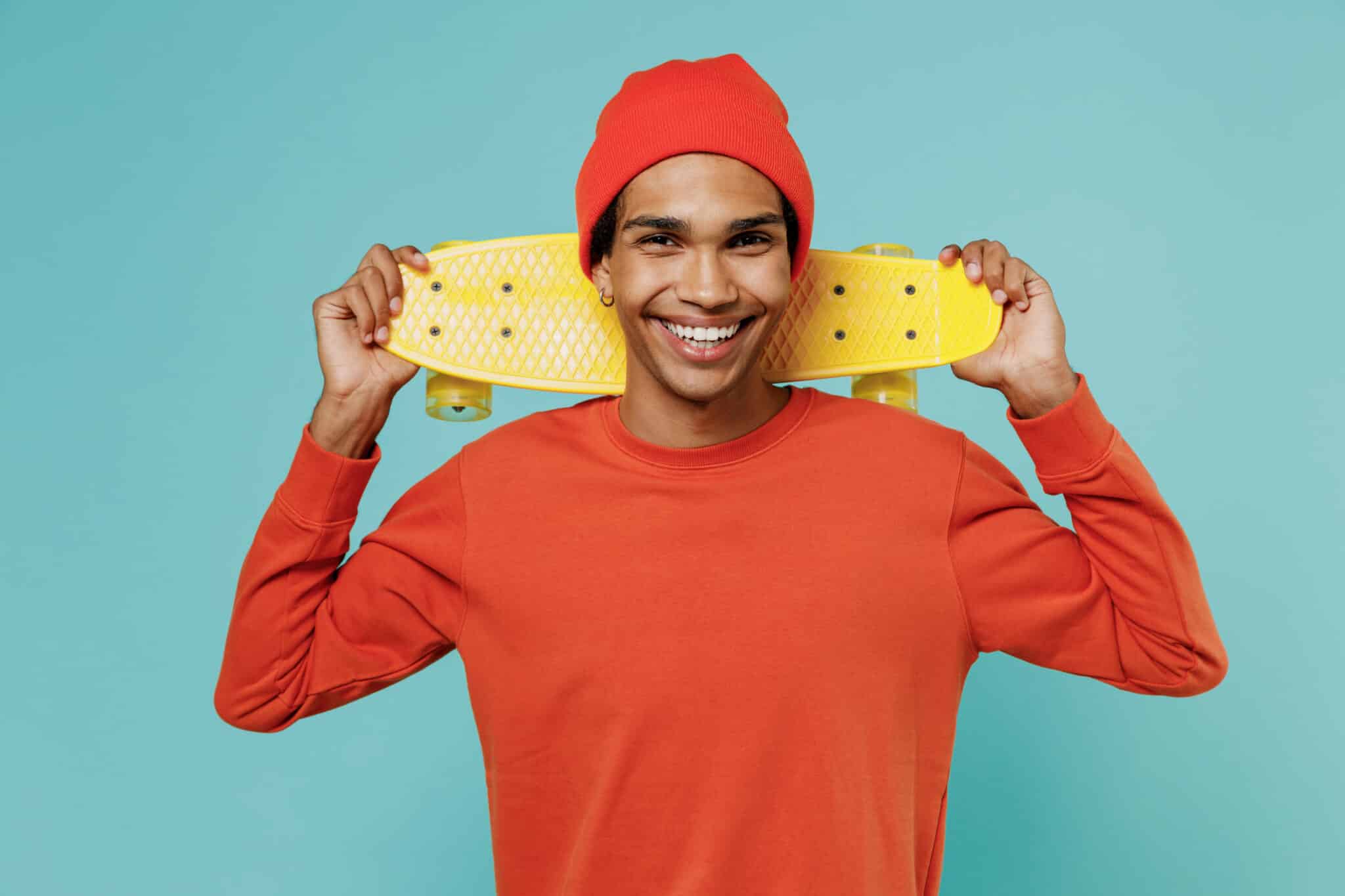Smiling teen with red hat and sweater carrying a skateboard.