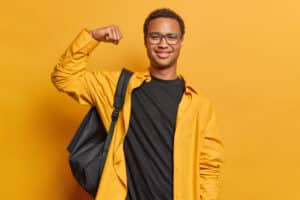 Cheerful young student happy for completing a personal goal. Wears a black t-shirt and backpack on yellow background. 