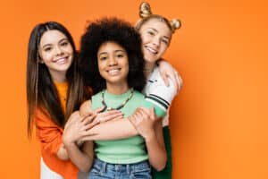 Three teen girls embracing and smiling at the camera on orange background. 